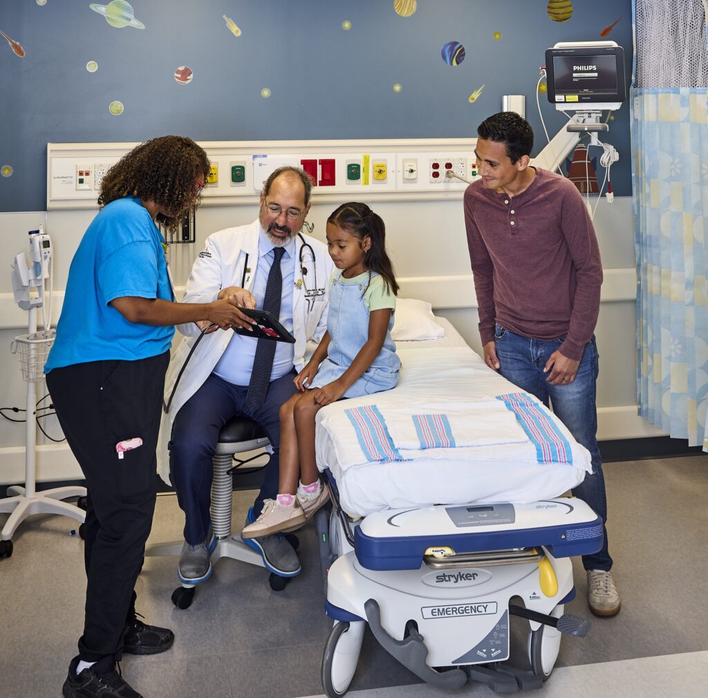 Doctor and nurse showing young patient sitting on hospital bed something on a tablet, while father looks on