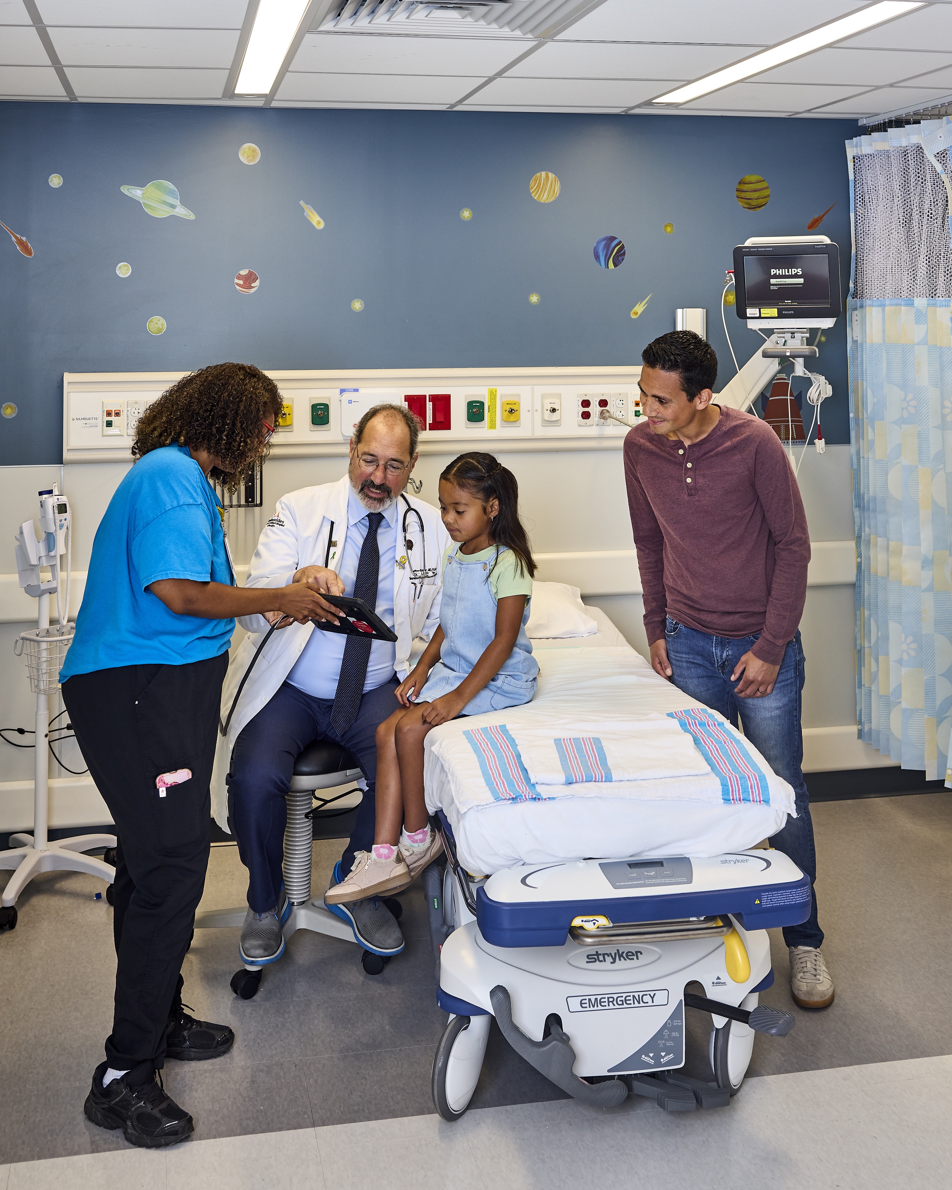 In treatment area, a doctor and nurse show a child something on a tablet while a parent looks on