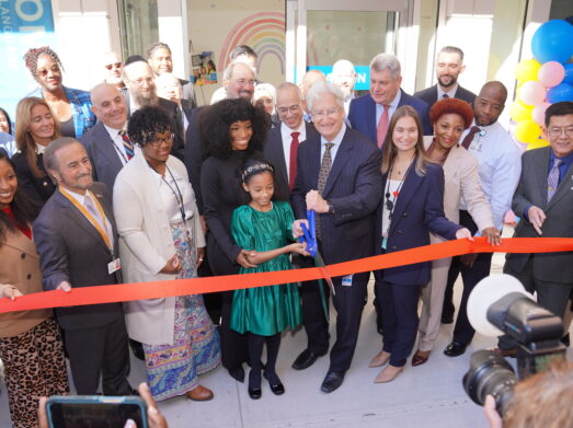 Child holds large scissors to cut ribbon, surrounded by large crowd