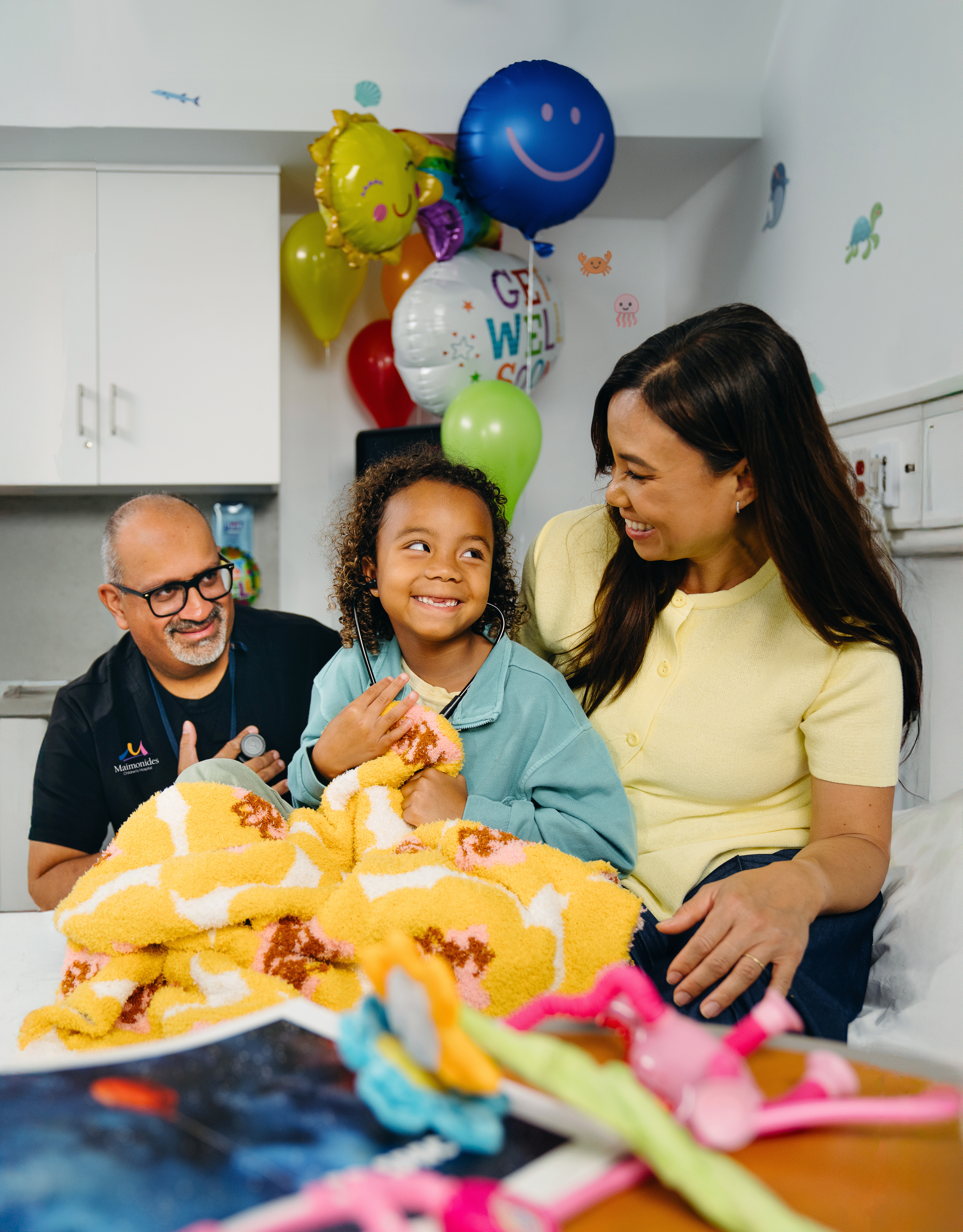 Doctor with smiling patient and mom, surrounded by colorful blankets and balloons in exam room